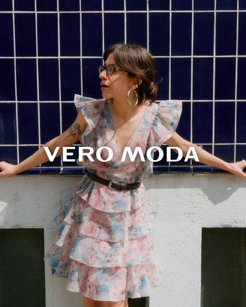 Una mujer con gafas y un vestido floreado con volantes se apoya en una pared blanca frente a un fondo de azulejos azules. La inscripción "VERO MODA" aparece en el centro de la imagen.