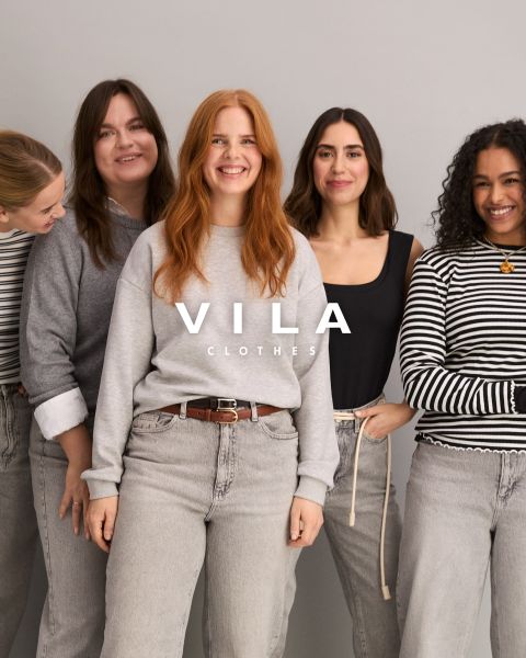 Cinco mujeres en ropa casual, incluyendo vaqueros y tops en colores neutros, están de pie juntas y sonriendo frente a un fondo sencillo. El texto superpuesto dice "Vila Clothes".