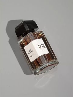 Un frasco de perfume de vidrio transparente con un tapón negro está sobre una superficie gris claro. En la etiqueta blanca se lee "Gris Chanel" y "bdk Parfums". El frasco proyecta una sombra suave.