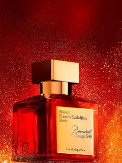 Un frasco del perfume Maison Francis Kurkdjian Paris Baccarat Rouge 540 con una tapa dorada se encuentra frente a un fondo rojo y dorado brillante.