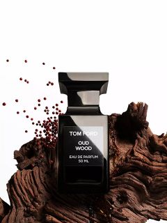 Un frasco negro del perfume Tom Ford Oud Wood está sobre un trozo de madera texturizada, cerca del cual hay pequeñas semillas marrones esparcidas, sobre un fondo blanco.