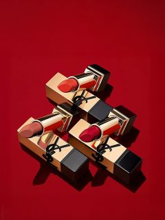 Tres labiales dorados y negros con diferentes tonos de rojo dispuestos diagonalmente sobre un fondo rojo liso. El empaque presenta un logo negro en la cubierta dorada.