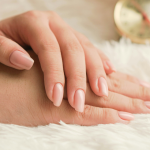 Zwei Hände mit sauber manikürten, hellrosa lackierten Nägeln ruhen auf einer weichen weißen Fläche, im Hintergrund ist ein unscharfes goldfarbenes Objekt zu sehen.