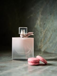 Un frasco de perfume rosa con un lazo está sobre una superficie de piedra junto a dos macarons rosas.