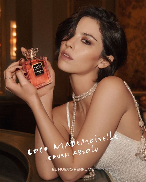 Mujer sosteniendo un frasco de perfume "Coco Mademoiselle", con accesorios de perlas, en un entorno elegante; el texto dice: "Coco Mademoiselle Crush Absolu, El Nuevo Perfume.".