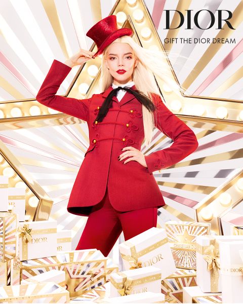 Una mujer con un llamativo traje rojo y sombrero de copa posa con confianza frente a una gran estrella iluminada, rodeada de cajas de regalo Dior blancas y doradas. El texto dice "DIOR REGALA EL SUEÑO DIOR".