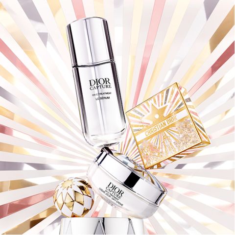 Dos productos de tratamiento Dior y un estuche de maquillaje dorado de Christian Dior se presentan sobre un fondo radiante con motivos de rayos de sol en tonos dorados, rosas y blancos, creando un efecto lujoso y luminoso.