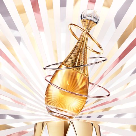 Un frasco de perfume dorado con elegantes anillos en espiral se encuentra frente a un fondo radiante de rayos dorados, rosados y blancos, creando una apariencia lujosa y glamurosa.
