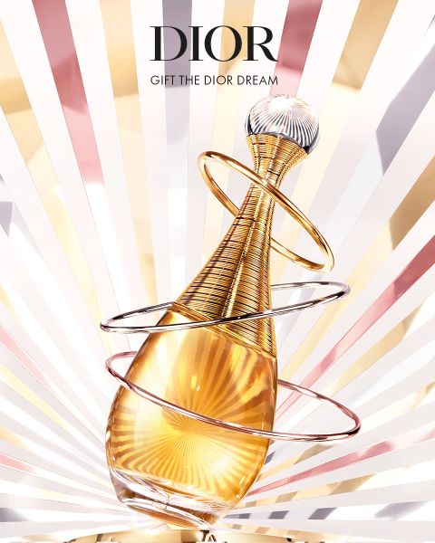 Un frasco dorado de perfume Dior con anillos en espiral frente a un brillante fondo de amanecer. El texto que lo acompaña dice: "DIOR" y "REGALA EL SUEÑO DIOR".