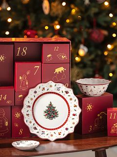 Una mesa festivamente puesta con platos y cuencos decorativos con motivos navideños se encuentra frente a un calendario de adviento rojo y un árbol de Navidad iluminado al fondo.