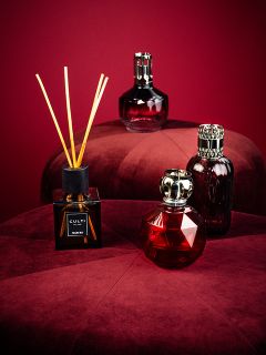 Cuatro elegantes frascos de perfume, incluido un difusor de varillas, están dispuestos sobre dos lujosos taburetes redondos de color burdeos frente a un fondo rojo oscuro a juego. Los frascos varían en forma y diseño y tienen un aspecto lujoso y sofisticado.