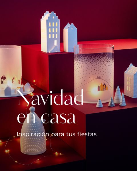 Casas de papel decorativas, árboles y candelabros con escenas invernales están dispuestos sobre superficies rojas iluminadas por luces cálidas. El texto en español dice: "Navidad en casa. Inspiración para tus fiestas.".