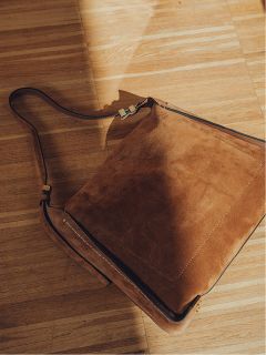 Un bolso bandolera marrón de ante con una correa larga está sobre un suelo de madera bañado por la cálida luz del sol, proyectando una suave sombra.