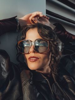 Una mujer con cabello castaño ondulado lleva gafas de sol grandes y una chaqueta de cuero oscura. Está apoyada contra una pared blanca, con las manos sobre la cabeza, y luce relajada y elegante.