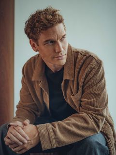 Un hombre con cabello castaño claro rizado está sentado en una casa y lleva una chaqueta de pana marrón sobre una camisa negra. Mira pensativo hacia un lado y tiene las manos entrelazadas relajadamente. El fondo está suavemente iluminado y es neutral.