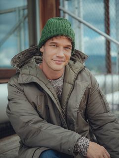 Un hombre con un gorro de lana verde, un suéter gris y una chaqueta de invierno verde oliva está sentado al aire libre en escalones de madera y sonríe suavemente a la cámara. En el fondo se ven una valla metálica y un edificio de cristal.