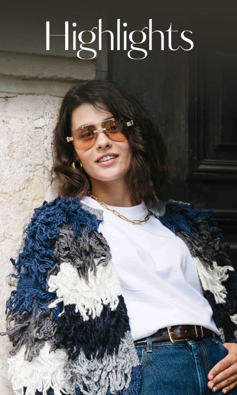 Una mujer con cabello castaño ondulado que lleva gafas de sol, un grueso suéter de punto azul y blanco, una camiseta blanca, vaqueros y un collar dorado está de pie junto a un edificio sonriendo. Encima de ella aparece la palabra "Highlights".