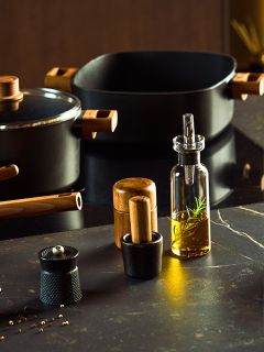Una encimera de cocina moderna muestra utensilios de cocina negros con mangos de madera, una botella de vidrio con aceite de romero, un molinillo de pimienta, un molinillo de sal y un pequeño mortero con maja, todo sobre una superficie oscura.