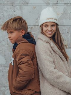 Un chico y una chica están de espaldas contra una pared de piedra clara. El chico lleva una chaqueta marrón y mira hacia un lado, mientras que la chica lleva un abrigo peludo y un gorro blanco y sonríe mientras mira hacia otro lado del chico.
