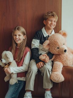Dos niños sonrientes están sentados uno al lado del otro, cada uno sosteniendo un peluche. La niña tiene el pelo largo y abraza un oso de peluche más pequeño, mientras que el niño sostiene un oso de peluche grande. Están sentados junto a una pared de madera y parecen felices y relajados.