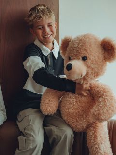 Un niño sonriente con cabello rubio está sentado en una casa abrazando a un gran oso de peluche beige. Lleva una camisa de manga larga blanca y negra y pantalones claros.