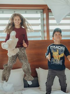 Dos niños, una niña con un peluche en la mano y un niño con una camiseta de Pikachu, saltan alegremente en una cama en una habitación luminosa con grandes ventanas al fondo.