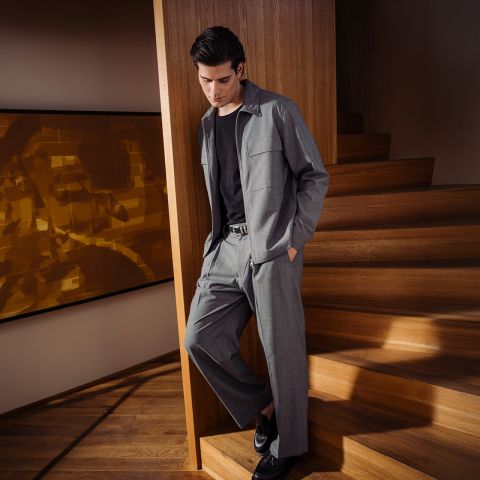 Un hombre con un traje gris, camisa negra y zapatos negros está de pie en una escalera de madera y se apoya casualmente en una pared en un interior moderno.