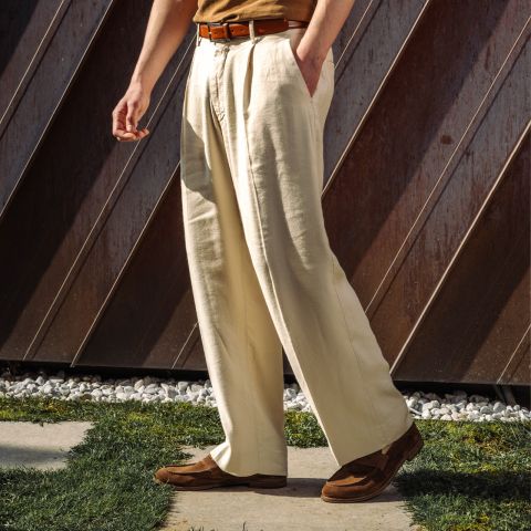 Una persona está de pie al aire libre y lleva unos pantalones de pinzas de color crema, un cinturón marrón, una camisa de color marrón claro y zapatos marrones, mientras la luz del sol proyecta sombras en el suelo.