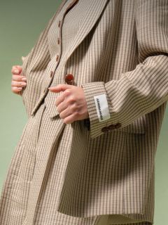 Una persona que lleva un traje a cuadros beige y marrón con una etiqueta blanca que dice "ABANDONABLE" en el puño, sujetando la solapa de la chaqueta frente a un fondo verde.