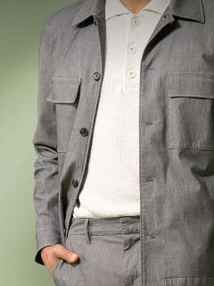 Una persona con una chaqueta de punto gris claro y pantalones a juego sobre un polo blanco texturizado, de pie con una mano en el bolsillo frente a un fondo verde tenue.