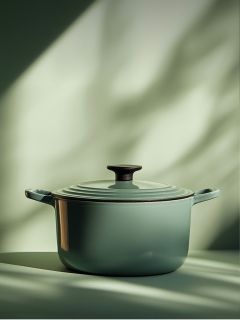 Una olla holandesa de hierro fundido esmaltada en verde con tapa y pomo negro se encuentra sobre una superficie iluminada por una suave luz natural con sombras borrosas al fondo.