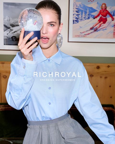 Una mujer con una camisa azul claro y pantalones grises sostiene una bola de nieve frente a un ojo. Posando en una habitación con paredes de madera y un póster de esquí, lleva pendientes grandes. El texto dice "RICHROYAL DRESSING SUPERWOMEN".