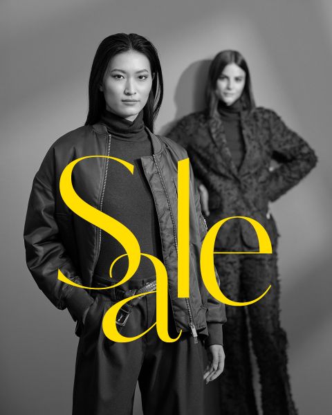 Dos mujeres posan con elegantes atuendos oscuros frente a un fondo gris con la palabra "Sale" en grandes letras amarillas que cruzan la imagen.