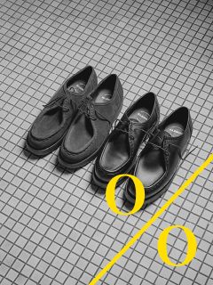 Dos pares de zapatos negros están uno al lado del otro sobre un suelo de baldosas, con dos círculos amarillos y dos líneas amarillas diagonales que atraviesan la parte inferior de la imagen.