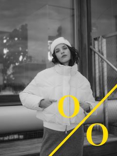 Una persona con una chaqueta de plumas blanca y un gorro posa al aire libre cerca de un edificio; una línea diagonal amarilla atraviesa la imagen con dos letras "O" amarillas a ambos lados. La foto está en blanco y negro.