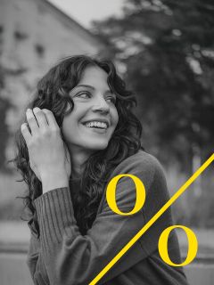 Una mujer con cabello rizado sonríe y mira hacia un lado. Lleva un jersey y está sentada al aire libre. Un gran signo de porcentaje amarillo se superpone a la foto en blanco y negro.