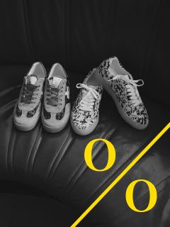 Una foto en blanco y negro de dos pares de zapatillas estampadas sobre un asiento de cuero negro. Una línea diagonal amarilla con dos letras "O" amarillas adorna la esquina inferior derecha de la imagen.