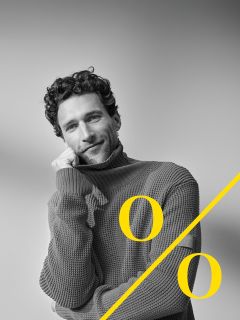 Un hombre con cabello rizado y barba lleva un grueso jersey de cuello alto y apoya su rostro sonriendo sobre su mano. Un gran signo de porcentaje amarillo se coloca diagonalmente sobre la imagen.