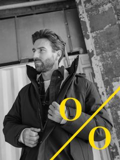 Un hombre con barba está de pie frente a un fondo industrial y se ajusta la chaqueta. La foto en blanco y negro muestra signos de porcentaje amarillos gruesos y una línea amarilla diagonal en la esquina inferior derecha.