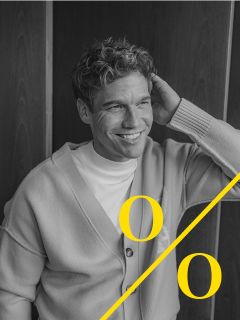 Un hombre con cabello rizado, que lleva una chaqueta de punto clara y una camisa blanca, está sentado sonriendo y sosteniendo una mano detrás de la cabeza. La imagen en blanco y negro muestra dos grandes "o" amarillas y una línea diagonal amarilla en la parte inferior.
