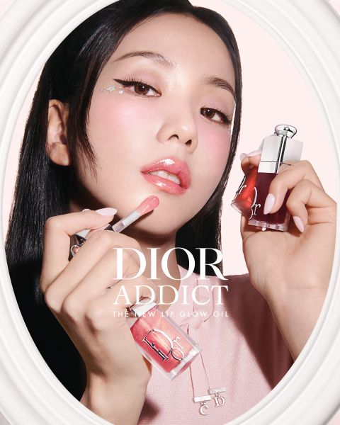 Una mujer se aplica brillo de labios rosa mientras sostiene una botella de Dior Addict Lip Glow Oil, con un maquillaje de color rosa pálido y una pegatina de estrella cerca de su ojo.
