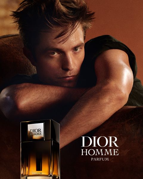 Un hombre se apoya en una superficie marrón junto a un frasco de Dior Homme Parfum, a su lado está el texto "DIOR HOMME PARFUM".