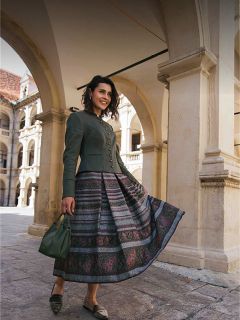 Una mujer con una falda estampada y una chaqueta verde está de pie bajo arcos de piedra, sonríe y sostiene un bolso verde en la mano.