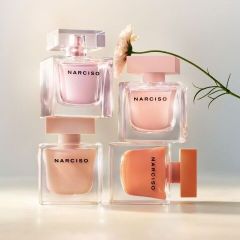 Cuatro frascos de perfume de la marca Narciso en tonos pastel están apilados uno encima del otro, y una única flor blanca se inclina desde la derecha.