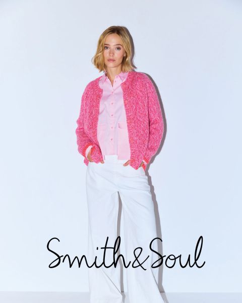 Una mujer con una camisa rosa y una chaqueta de punto con pantalones blancos posa delante de un fondo blanco. El texto dice "Smith & Soul" en la parte inferior.