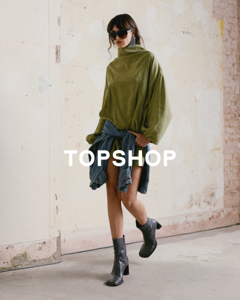 Una mujer con una camiseta verde de gran tamaño, una chaqueta vaquera atada a la cintura, botas negras y gafas de sol está de pie frente a una pared desgastada. El texto "TOPSHOP" está superpuesto.