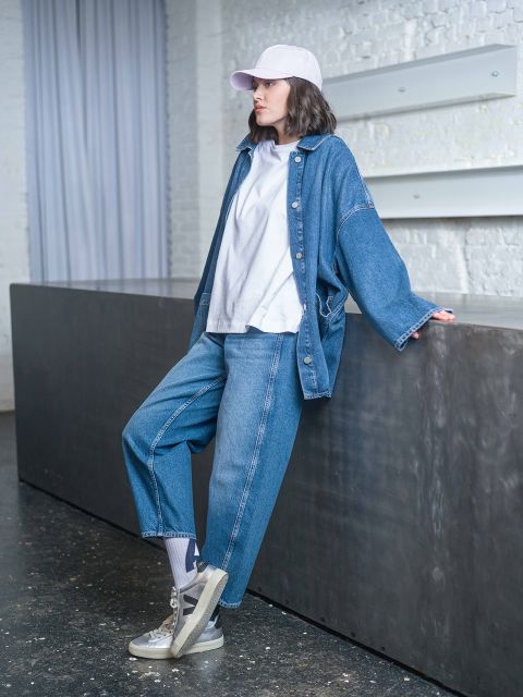 Una persona con una chaqueta vaquera, una camiseta blanca y una gorra blanca está apoyada en una barra oscura en un interior moderno.