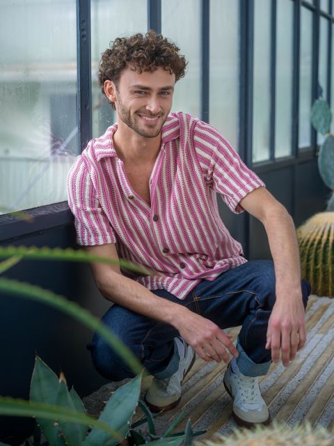 Un hombre con pelo rizado, con una camisa de rayas rosas y vaqueros, está en cuclillas en una casa cerca de plantas y sonríe a la cámara.