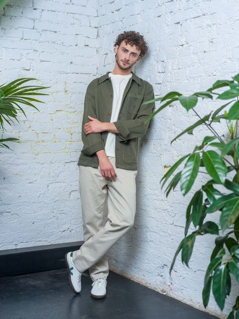 Un hombre con cabello rizado, con una chaqueta verde, camisa blanca y pantalones beige, está apoyado contra una pared de ladrillo blanco, rodeado de plantas verdes.