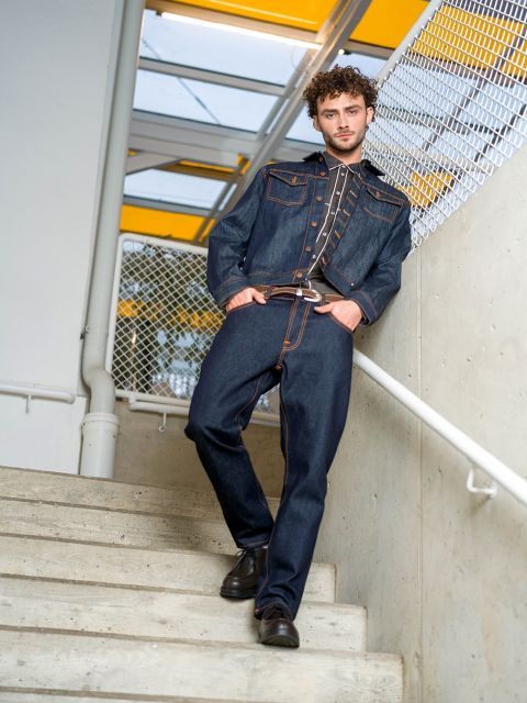 Un hombre con chaqueta vaquera oscura y vaqueros está de pie en una escalera, con un pie más arriba, y se sujeta a la barandilla, detrás de él una valla de tela metálica y una claraboya amarilla.